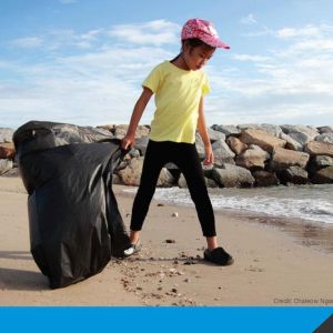 A child looks down at a sandy beach while holding a trash bag