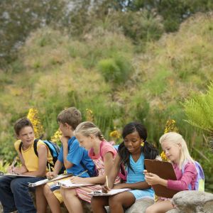 five elementary students writing on clipboards and sitting on rock wall in front of green shrubbery
