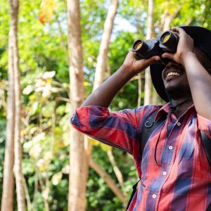 Smiling person in red flannel and fishing hat looking through pair of binoculars in wooded area