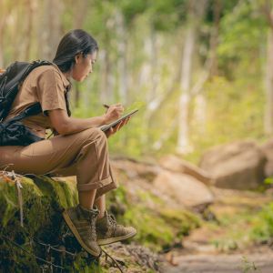 Person wearing backpack, sitting on mossy stones in forest to take some notes.