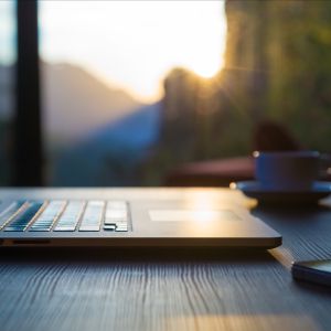 Laptop, mug, and phone lay on black wood table as sunlight shines through large windows