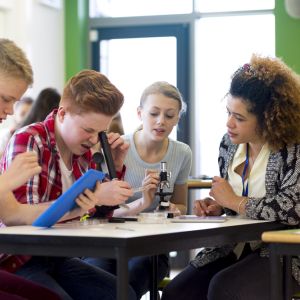 Students sitting with teacher in a lesson. They are using microscopes and a digital tablet.