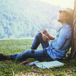 man sitting against tree writing in notebook and looking up at trees on hillside