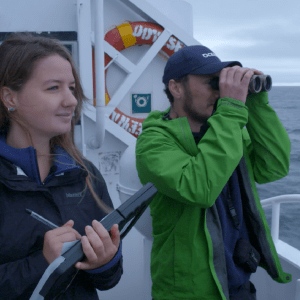 Two researchers looking at the ocean from a boat. One has a binocular while the other is taking notes.