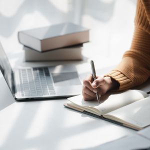 Person writing in a notebook with laptop and books to the side
