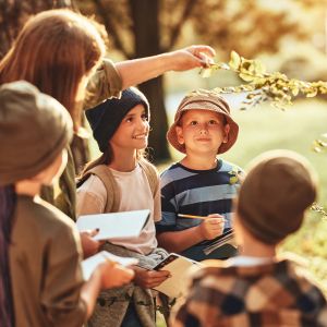 A group of children are crowded around a central adult who is reaching out to touch the branch of a leafy plant