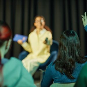 Teenage girl with brown hair sitting on stage holding tablet and remote, speaking to audience while teenage girl with dark hair raising hand in foreground, conference setting