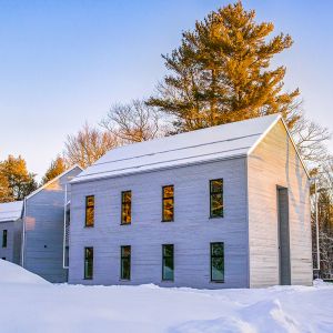 A light-gray building with an A-frame roof stands prominently in morning sunlight. A layer of snow sits on the roof and deep snow is on the ground. Behind the building are leafless trees and conifers, lit brightly by yellow morning sunlight. Above the trees is blue sky.