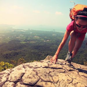 Hiker climbing rock on mountain peak. Lush green landscape below.