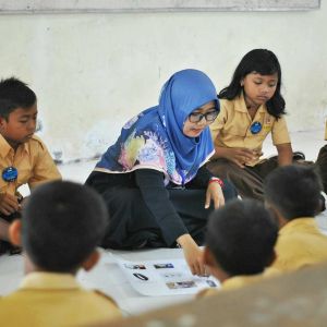 A group of children and an adult, who appears to be a teacher, are sitting on the floor in a classroom setting.