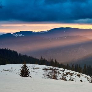 Photo of a snowy hill, forest silhouette in the back, and rows of mountain ranges higlighted by a few rays of sunlight peeking out under a dark blue cloudy sky