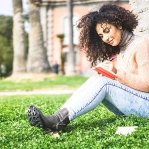 Person sitting on grass against a tree, writing in small journal.