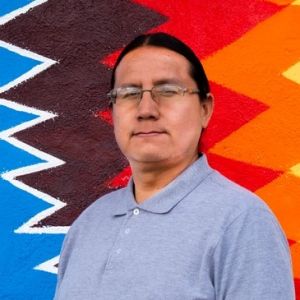 Profile photo of a man wearing glasses, hair pulled back, and staring straight at the camera. He stands in front of a colorful, geometric backdrop
