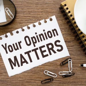 "Your opinion matters" written on notebook paper, sitting on wooden table next to notebook, coffee, paper clips