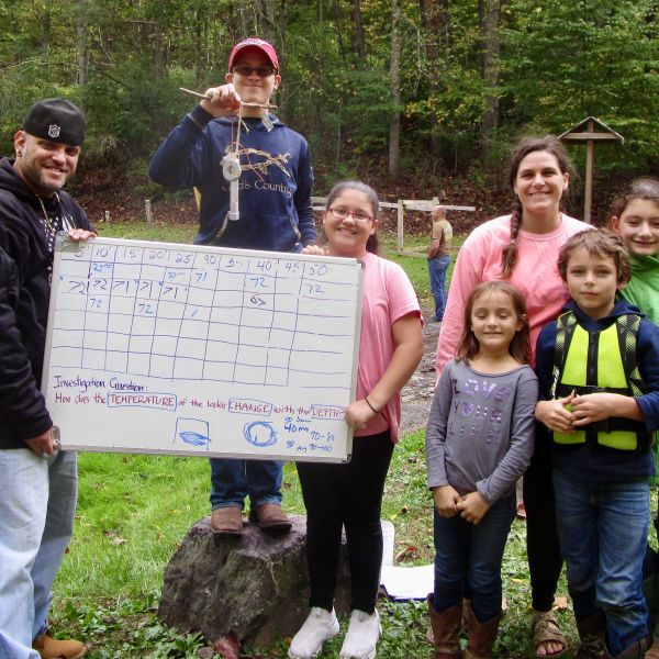 A photo of a family holding a whiteboard and thermometer in a forest clearing.