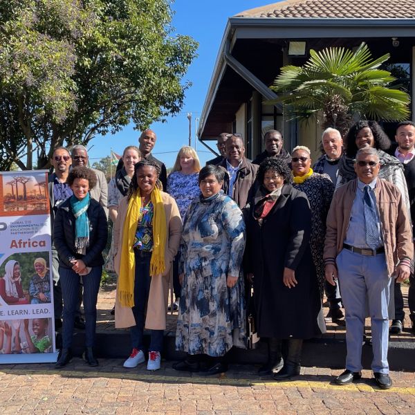 A group of EE Leaders from around the world smile for a picture outside. Trees and building in background.