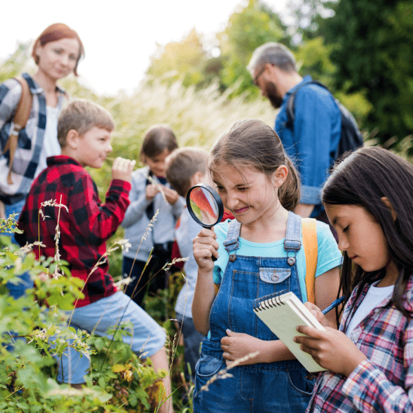Group of students outside using a magnifying glass and a notebook to record what they see in a shrub