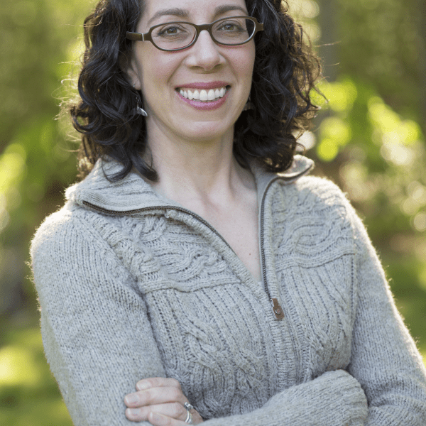 image of Jeanine Silversmith with medium length dark curly hair, glasses, smiling, and wearing a light gray sweater, standing in front of leafy green trees