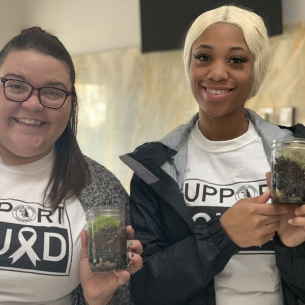 Two women smile at the camera while each holding a mason jar filled with rocks, soil, and green vegetation.