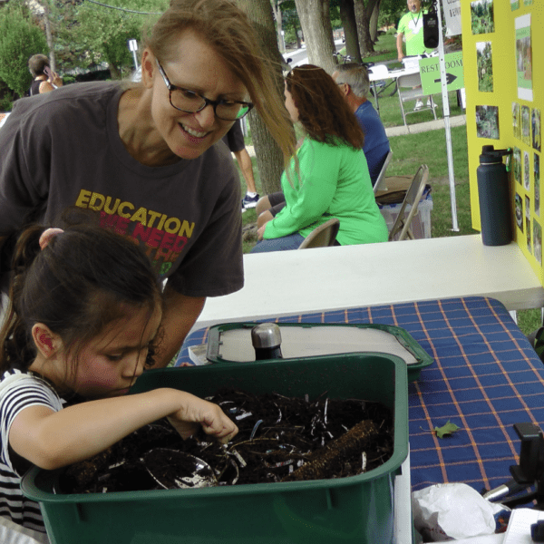 CEE-Change Fellow Melanie Schikore engages children at the farmer's market. Melanie is smiling while one child is focused and picking up worms from soil in the green worm bin.