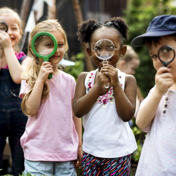 four young children holding magnifying glasses outside in green space