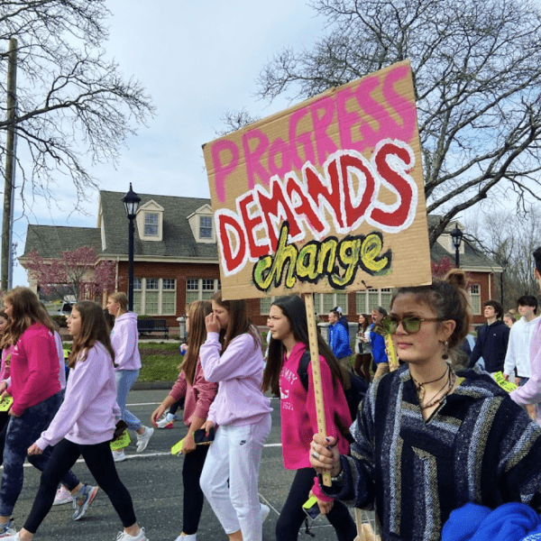 Dozens of teenage students walk through the street. One holds a sign saying "Progress demands change."