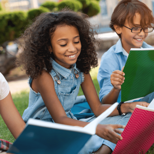 Three students sitting outside on a grassy area. Each young student holds open a notebook.