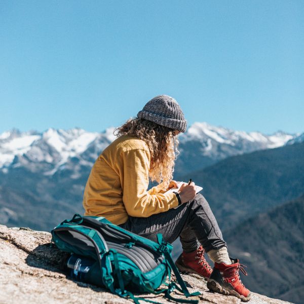 person with long curly hair wearing grey knit cap, yellow top, grey pants, red hiking shoes, sits on mountain top writing, blue backpack sits behind them