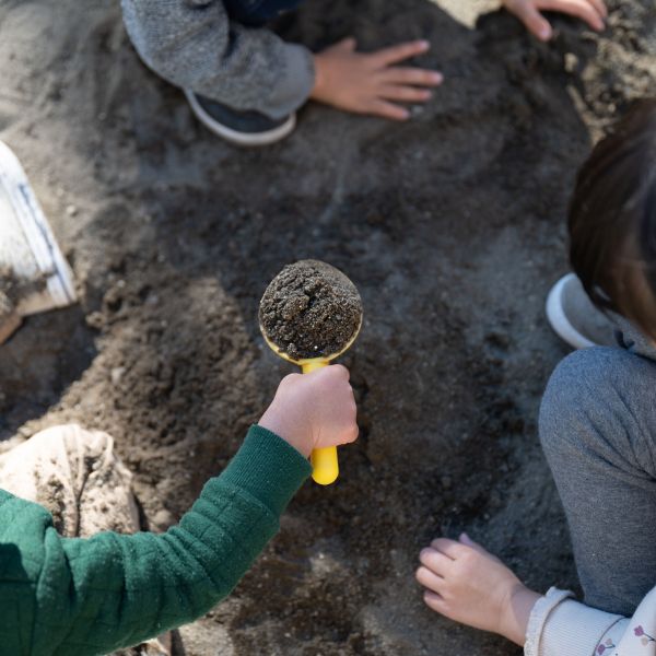 Three kids in a sandbox where one of the kids holds a scooper filled with sand