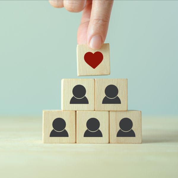 A hand places a wooden block with a red heart on it atop a pyramid made with blocks, each with a human shape on it.