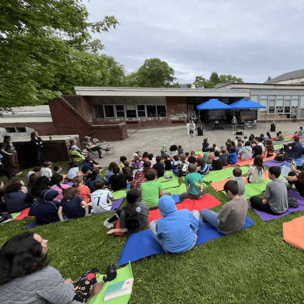 Large group of students seated on a down-sloping lawn facing a paved stage in front of school buildings