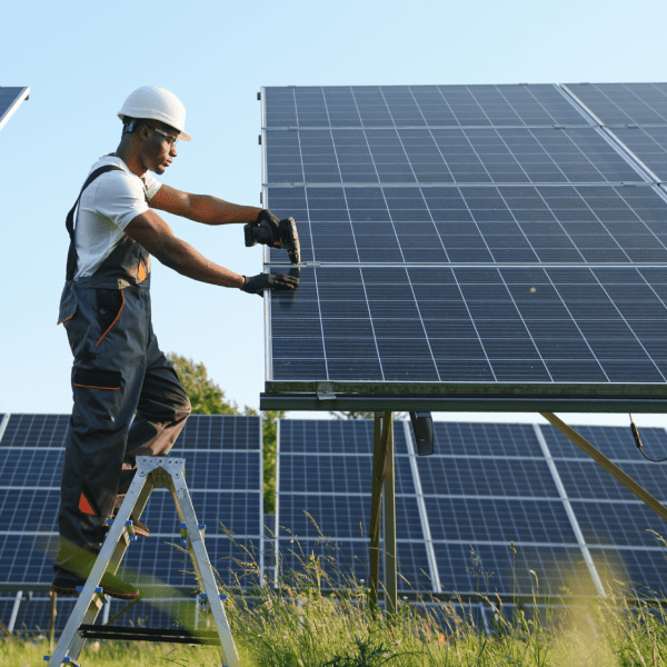 A person in a white hard hat stands between two solar panels, working on the one to the right