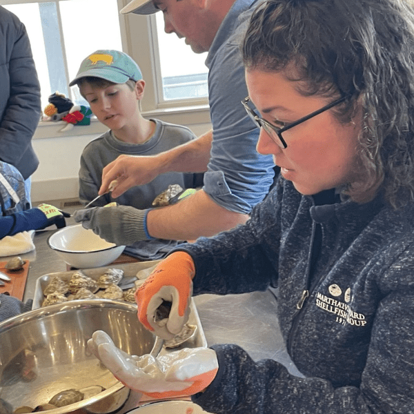 A curly-haired woman wearing glasses and orange gloves holds an oyster. In the background are two young boys watching as an adult shucks an oyster. They are all indoors.