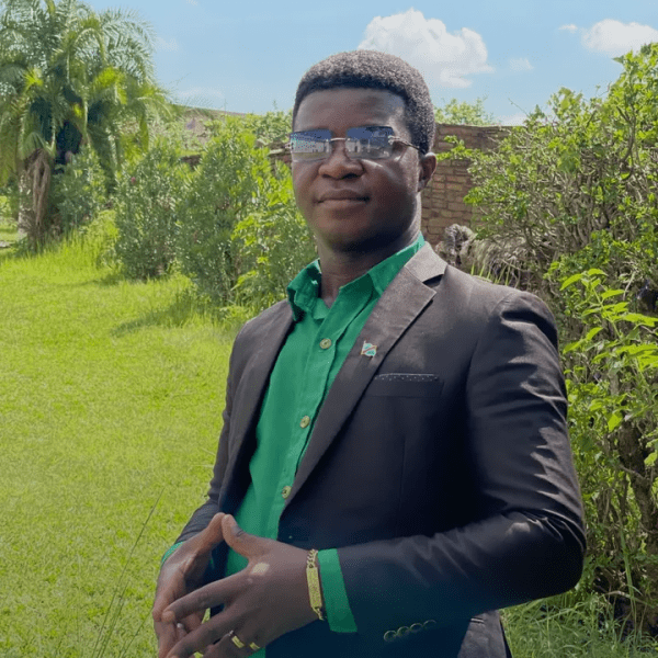 Kristian Katele in a green button up and black blazer stands outside under a sunny scky