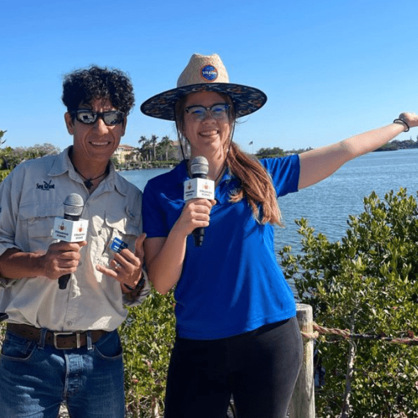Armando and Caroline stand next to each other in front of a clear blue sky, water, and treeline; they both hold microphones
