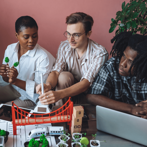 Four people sit at a table with miniatures of a bridge, trees, buses, a wind turbine, as well as papers and a laptop