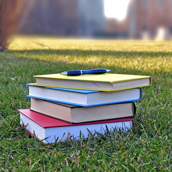 Four books stacked on a grass lawn with a pen on top of them; the background is a blurred university campus