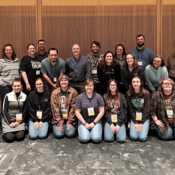 Group photo of people kneeling in front of people standing, all wearing nametags around their necks, on a carpeted floor with a wood paneled wall behind them