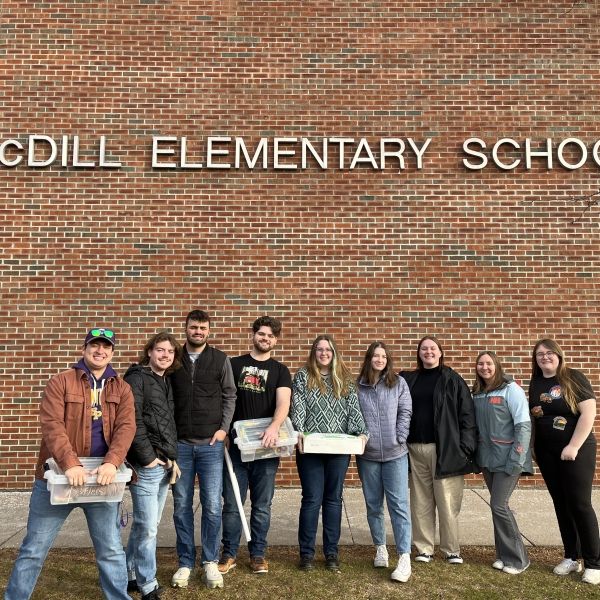UWSP Environmental Education and Interpretation students stand in front of a red brick wall with the words "McDILL ELEMENTARY SCHOOL