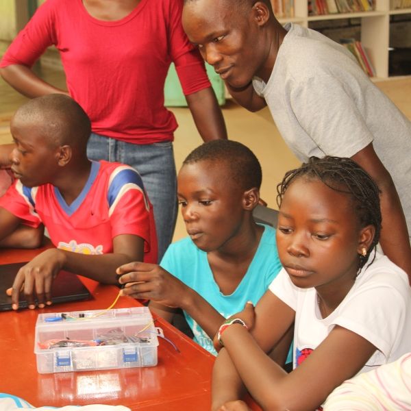An Arduino class, six students sit around a table with electronic wires in the middle, three adults stand behind them