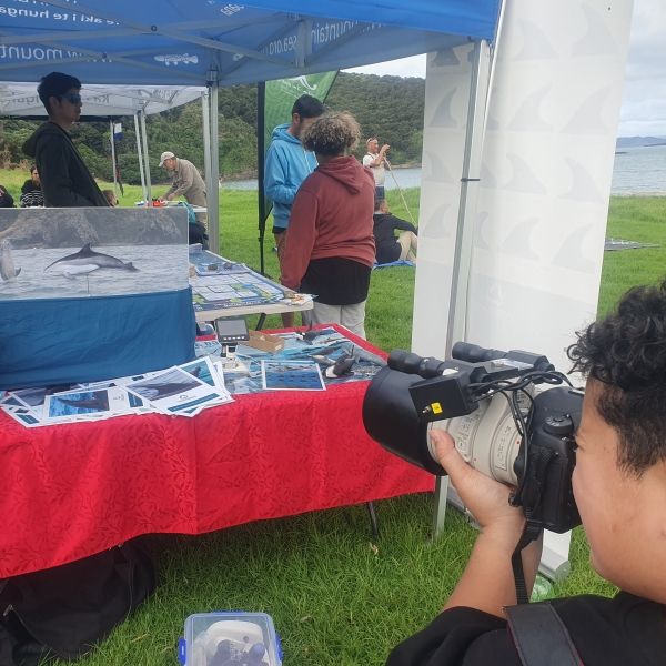 A person holds a camera pointing away from the viewer and toward a covered booth with information about dolphins, people are standing around it