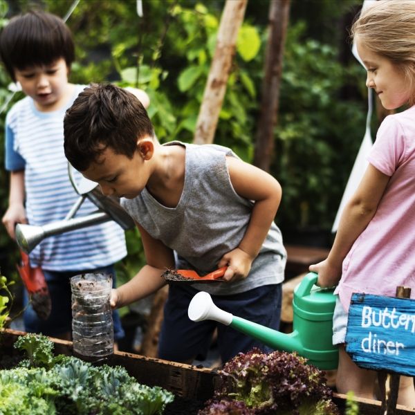 Three children around a boarded garden bed, two holding watering cans and one holding a plastic bottle with dirt