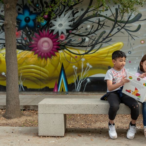 A first-grade boy reads a book to his kindergarten friend while sitting outside.