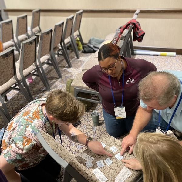 Four conference attendees kneel around a chair in a meeting room, collaborating on a hands-on activity by arranging and discussing cards spread across the seat.