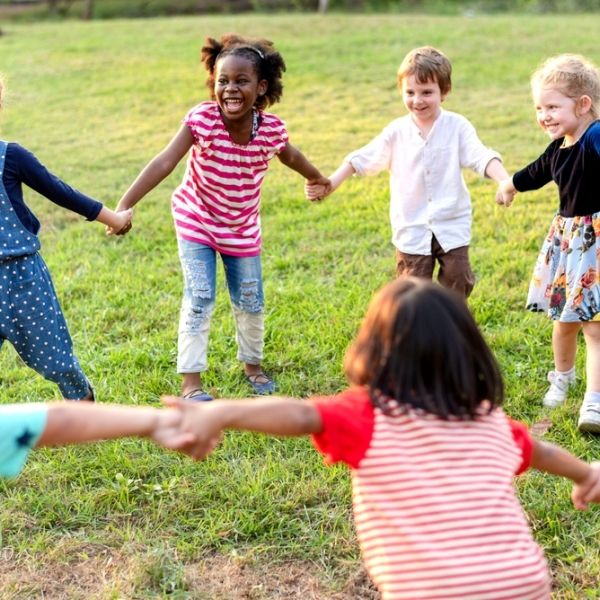 Young children smile and hold hands in a circle on green grass.