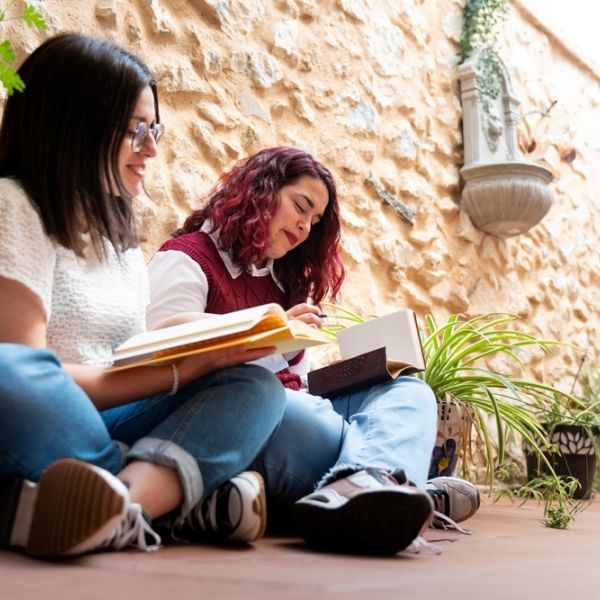 Two women site side by side reading books outside against a wall.